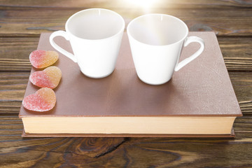 Two cups on a book with candy in the form of a heart on a wooden table background.