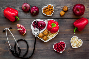 Products good for heart and blood vessels. Vegetables, fruits, nuts in heart shaped bowl near stethoscope on dark wooden background top view