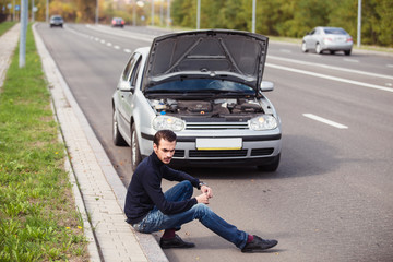 A young man sitting on the road in front of his silver car and thinking how to repair it