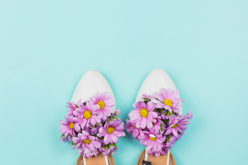 White flat female shoes filled with pink daisies and wildflowers on pastel blue background. Concept of summer or spring, copyspace