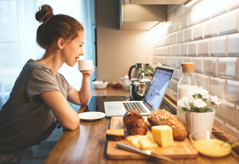 woman in pajamas is having breakfast at computer in  kitchen. - Powered by Adobe