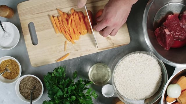 Close up hands of male cook cutting carrot into sticks on wooden cutting board with other ingredients placed around it, top view
