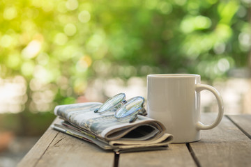 Cup of hot coffee, reading glasses and newspaper on wooden table in the garden morning