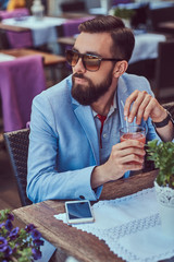 Portrait of a fashionable bearded male with a stylish haircut, holds a glass of a cappuccino, sitting in a cafe outdoors.