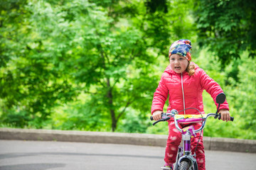 Fototapeta na wymiar A little girl in a pink jacket is riding a bicycle. Emotional child riding a bicycle on a background of a green forest