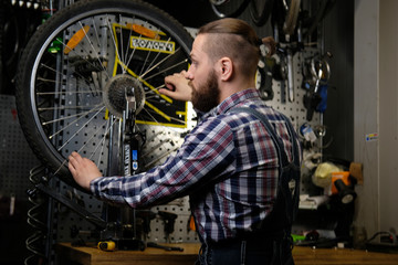 Handsome stylish male wearing a flannel shirt and jeans coverall, working with a bicycle wheel in a repair shop.