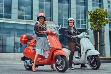 Fototapeta na wymiar Attractive romantic couple, a handsome man and sexy female, standing with two retro Italian scooters against a skyscraper.