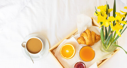 Fototapeta na wymiar Morning breakfast in bed wooden tray with a cup of coffee croissant orange juice fresh orange jam bouquet of flowers daffodils. Top view Morning at Hotel Background Concept Interior Copy Space