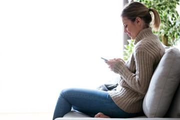 Pretty young woman using her mobile phone while sitting on sofa at home.