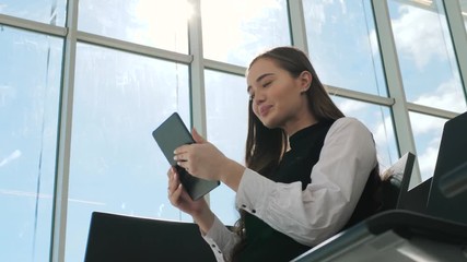 Young female passenger at the airport, using her tablet computer while waiting for her flight, smiling, sunnu day, terminal raiyway station bus transportation, business woman student