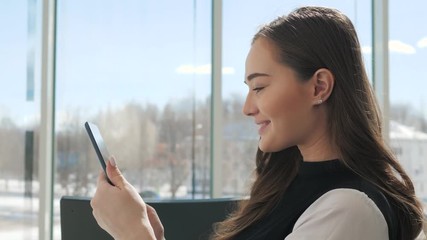 Young female passenger at the airport, using her tablet computer while waiting for her flight, smiling, sunnu day, terminal raiyway station bus transportation, business woman student