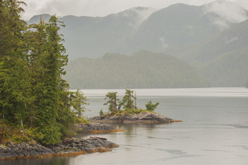 Kleine bewaldete Inselchen. Die Inside Passage in Westkanada, tr&uuml;bes Wetter, verhangene Berge im Hintergrund
