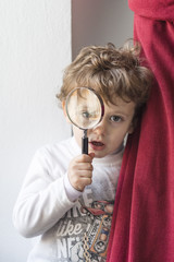 Child hiding behind a red curtain looking with a magnifying glass