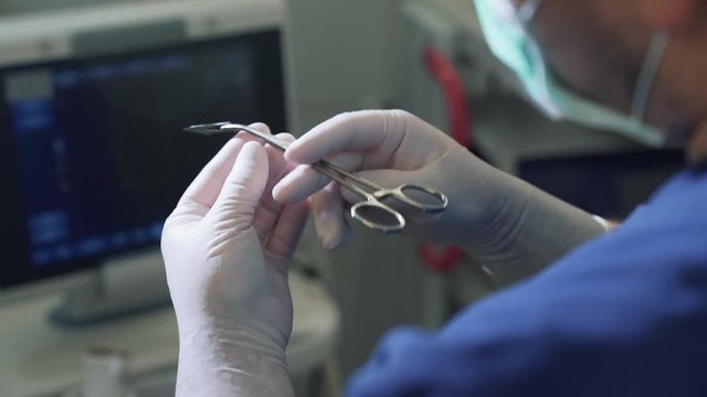 Personal Perspective, The Doctor In Sterile Gloves Holds The Scalpel Blade In The Clamp Before The Operation. POV Close Up Medical Instruments In Hands, View From The Back, Equipment In A Clinic And