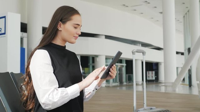 Young female passenger at the airport, using her tablet computer while waiting for her flight, smiling, sunnu day, terminal raiyway station bus transportation, business woman student - Powered by Adobe