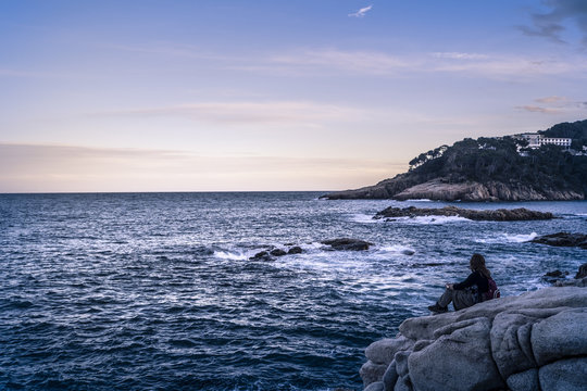 Woman looking at sea while sitting on rocks against sky during sunset