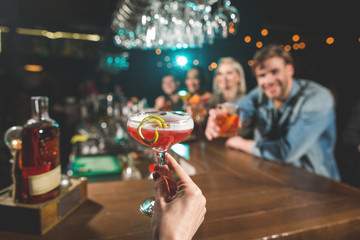 Let us drink. Close up female hand holding appetizing cocktail. Comrades sitting opposite her. Toast for friendship concept