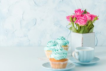 Blue pastel horisontal banner with decorated cupcakes, cup of coffe with milk and bouquet of pink roses.