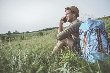 Naklejka na ściany i meble In touch. Side view of positive hiker is sitting on the grass while talking on smartphone with his friend. Backpack is lying near. Copy space in the left side