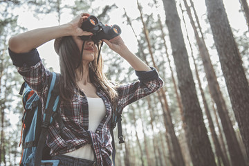 Wild place. Low angle of modern girl with backpack is looking through binoculars while standing in the forest. Copy space in the right side