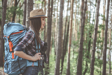 Naklejka premium Action adventure. Side view profile of pleasant optimistic young woman wearing hat is standing in forest with rucksack and binoculars. She is looking ahead with slight smile. Copy space in right side