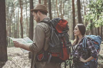Our location. Side view of young pleasant man and woman are standing with backpack in the forest. Guy is holding map and looking at it with concentration