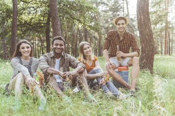 joyous men and women reposing on grass with bottles of beer in hands. They are looking at camera with smile. Portrait