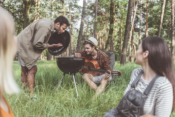 Fototapeta premium Two men opening barbecue cover and smelling smoke with pleasure. Smiling women sitting and looking at them
