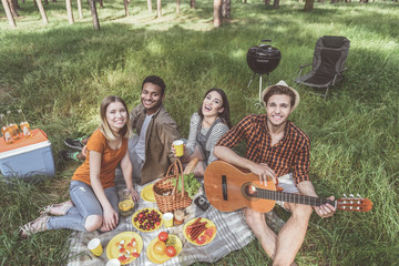 Portrait of four enjoyed people sitting on blanket full of food. They are staring at camera and laughing. Man holding guitar