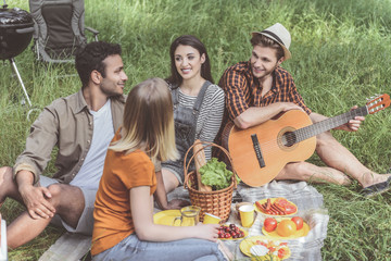 Satisfied friends having picnic with guitar on grass. They are talking and laughing
