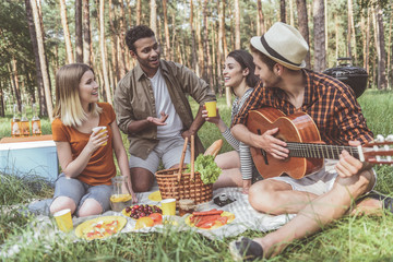 Young delighted men and women relaxing on blanket with food on it. They are speaking to each other and laughing. Guy playing guitar with joy