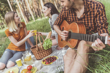 Man and women having picnic on rug. Guy sitting with musical instrument while girls clinking disposable cups and laughing