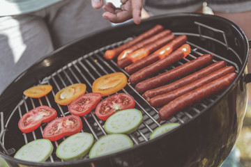barbecue grate with vegetable and sausages on it. Friends standing nearby. Close up