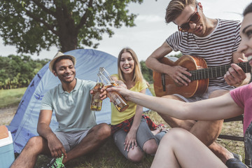 Cheers. Joyous man and women relaxing on grass and clinking bottles of beer. Focus on calm male...