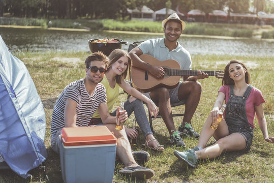 Four happy friends with beer and guitar in hands sitting at a lake. They are looking at camera with satisfaction