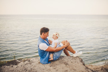 Family summer vacation in nature. father and little daughter sit on sandy cliff for one year with high view of the sea. A man is holding child in his arms. Emotion of happiness, joy and parental love