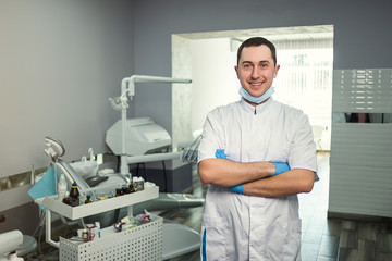 Naklejka na ściany i meble Male dentist standing with his hands crossed over medical office background. Healthcare, profession, stomatology and medicine concept