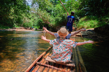 Boys (8 years old) raising one's hands floating along tropical river on bamboo raft in jungle. Concept traveling with children.