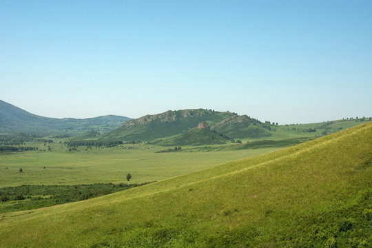 Minimalistic green landscape with diagonal side of hill on foreground, beautiful rocky hills and clear blue sky on background. Unique hill in hat form in distance.
