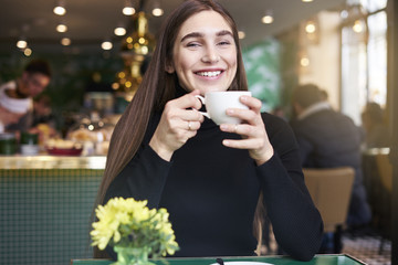 Young woman with long hair smiling, drinking cup of coffee in hands having rest in cafe near window