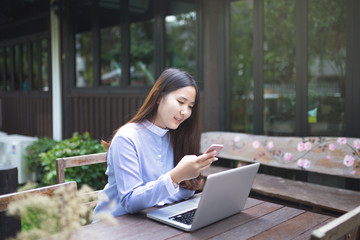 Women sitting and using smartphone and laptop on the table and she is feeling happiness at garden or cafe shop.