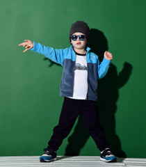 Preschool boy kid standing in blue zip-up sweatshirt and gray hat in shorts and sunglasses happy pointing hand 