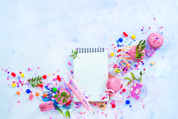 Planning a party concept with confetti, pink macarons, candies and an open notepad with blank pages. Creative celebration flat lay with copy space.