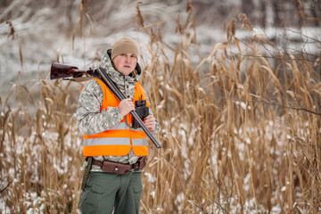 Male hunter in camouflage, armed with a rifle, standing in a snowy winter forest with duck prey