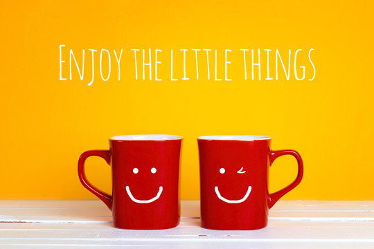 Two red coffee mugs with a smiling faces on a yellow background with the phrase Enjoy the little things.