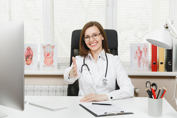 Young smiling female doctor standing with outstretched hand for greeting, sitting at desk with medical documents in light office in hospital. Woman in medical gown, stethoscope in consulting room.