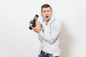 Young handsome surprised man student in t-shirt and light sweatshirt with hood with headphones holds opened retro camera and shows it to camera isolated on white background. Concept of photography