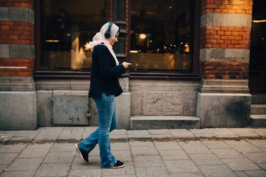 Full length side view of teenage girl walking on sidewalk listening to headphones while using smart phone in city
