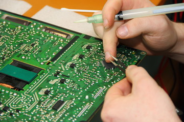 The hands of the master hold a tweezer with a chip and a syringe with flux solder over the PCB
