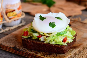 Vegetable salad and poached egg sandwich. Poached egg on rye bread slice with fresh cabbage, cucumber, red pepper and parsley. Healthy egg breakfast. Wooden background. Rustic style. Closeup